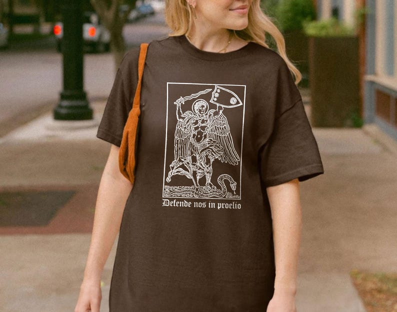 May include: A dark brown t-shirt featuring a white graphic of an angel with a sword and shield, and the text "Defende nos in proelio" in a rectangular frame. The shirt has short sleeves and a relaxed fit.