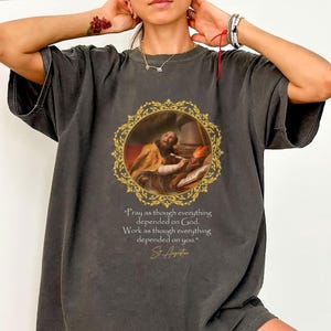 Op de afbeelding: Donkergrijs T-shirt met een Renaissance-schilderij in een gouden lijst. Het shirt toont de quote: "Bid alsof alles van God afhangt. Werk alsof alles van jou afhangt." toegeschreven aan St. Augustinus.