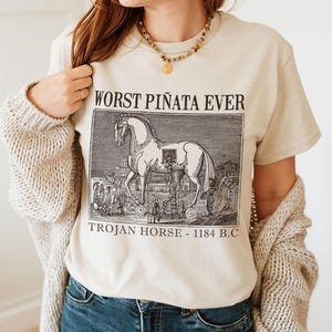 May include: Beige t-shirt with a black and white illustration of a Trojan horse and the text "WORST PI&Ntilde;ATA EVER TROJAN HORSE - 1184 B.C." The shirt is made of a soft material.