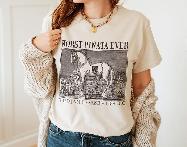 May include: Beige t-shirt with a black and white illustration of a Trojan horse and the text "WORST PI&Ntilde;ATA EVER TROJAN HORSE - 1184 B.C." The shirt is made of a soft material.