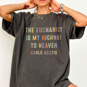 May include: Dark gray t-shirt with the text "THE EUCHARIST IS MY HIGHWAY TO HEAVEN CARLO ACUTIS" in colorful block letters. The shirt has a relaxed fit and short sleeves. The text is in a rainbow of colors.