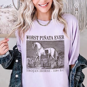 May include: A light purple t-shirt with the text "WORST PI&Ntilde;ATA EVER" above a black and white illustration of the Trojan Horse. The text "TROJAN HORSE - 1184 B.C." is below the illustration. The shirt is worn with a denim jacket.