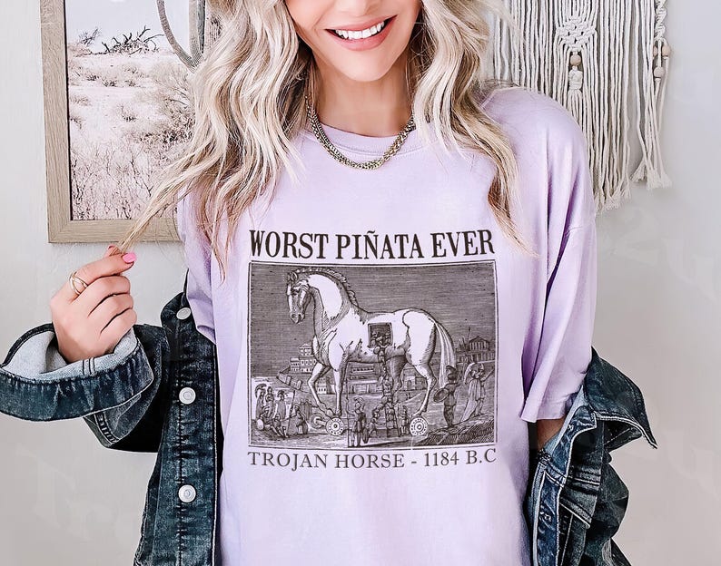 May include: A light purple t-shirt with the text "WORST PI&Ntilde;ATA EVER" above a black and white illustration of the Trojan Horse. The text "TROJAN HORSE - 1184 B.C." is below the illustration. The shirt is worn with a denim jacket.