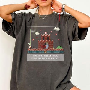 May include: Dark gray t-shirt featuring a pixelated castle scene with the text "HAIL MARY FULL OF GRACE, PUNCH THE DEVIL IN THE FACE!". The graphic includes a red cross, clouds, and a green level bar.