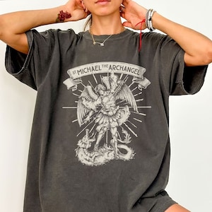 May include: A dark gray oversized t-shirt with a graphic print of an angel with a sword fighting a dragon. The text "St. Michael the Archangel" is printed above the graphic.