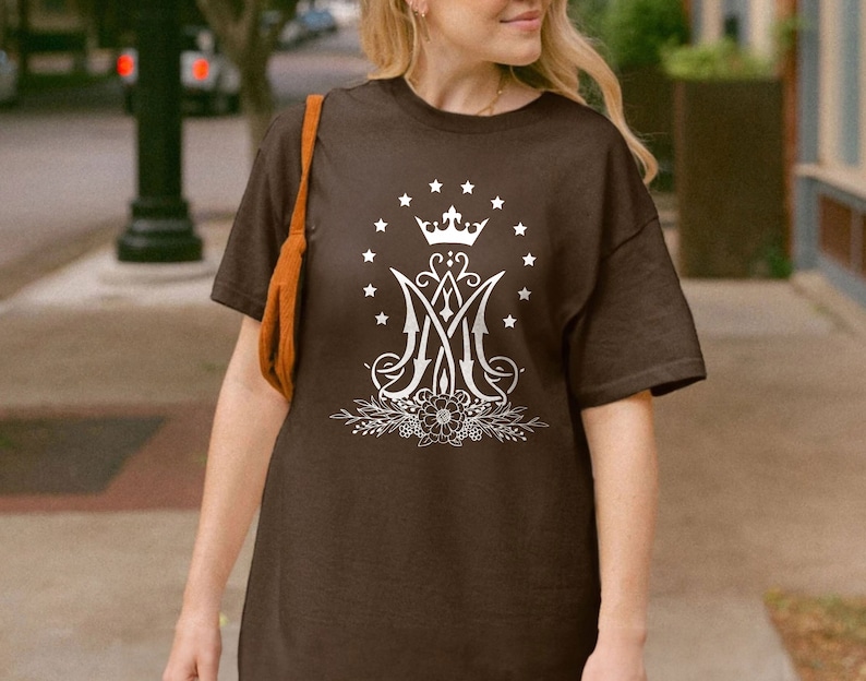 May include: A brown t-shirt featuring a white graphic of a crown, stars, and the letters "M" and "A" intertwined. The graphic is surrounded by a floral design.