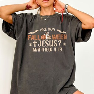 Può includere: Una maglietta nera con la scritta "Are you Fall-oween + Jesus? Matthew 4:19" stampata su di essa. Il testo è circondato da immagini di zucche e pipistrelli.