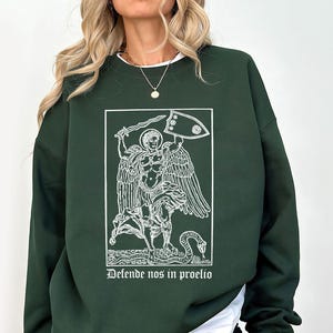 May include: Dark green sweatshirt featuring a white graphic of a winged figure holding a sword and shield, with the text "Defende nos in proelio" below. The design is framed within a rectangle.