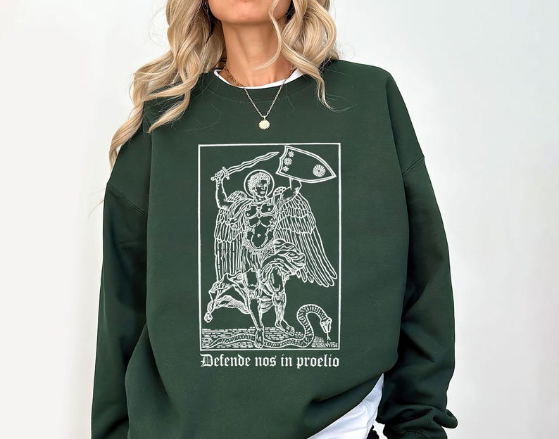 May include: Dark green sweatshirt featuring a white graphic of a winged figure holding a sword and shield, with the text "Defende nos in proelio" below. The design is framed within a rectangle.