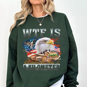 May include: Dark green sweatshirt featuring a graphic of an American bald eagle, the American flag, and the text "WTF IS A KILOMETER". The design includes a tank and a burger. The sweatshirt is a casual style.