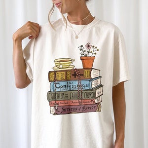 May include: A white t-shirt with a graphic of a stack of books, a cup of tea, and a potted plant. The books are labeled "HOLY BIBLE", "the Confessions", "CATECHISM of the CATHOLIC CHURCH", "The Imitation of CHRIST", and "DOUAY RHEIMS".