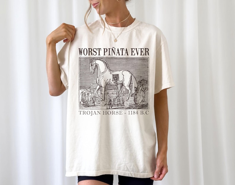 May include: Cream-colored t-shirt featuring a black and white illustration of the Trojan Horse with the text "WORST PI&Ntilde;ATA EVER" and "TROJAN HORSE - 1184 B.C." printed on the front. The shirt has short sleeves and a relaxed fit.