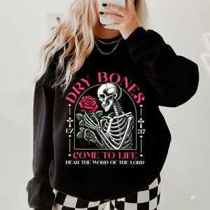 May include: Black sweatshirt with a graphic of a skeleton holding a red rose. The graphic is surrounded by a black frame with the text "DRY BONES" in pink letters and "COME TO LIFE - HEAR THE WORD OF THE LORD" in smaller pink letters.