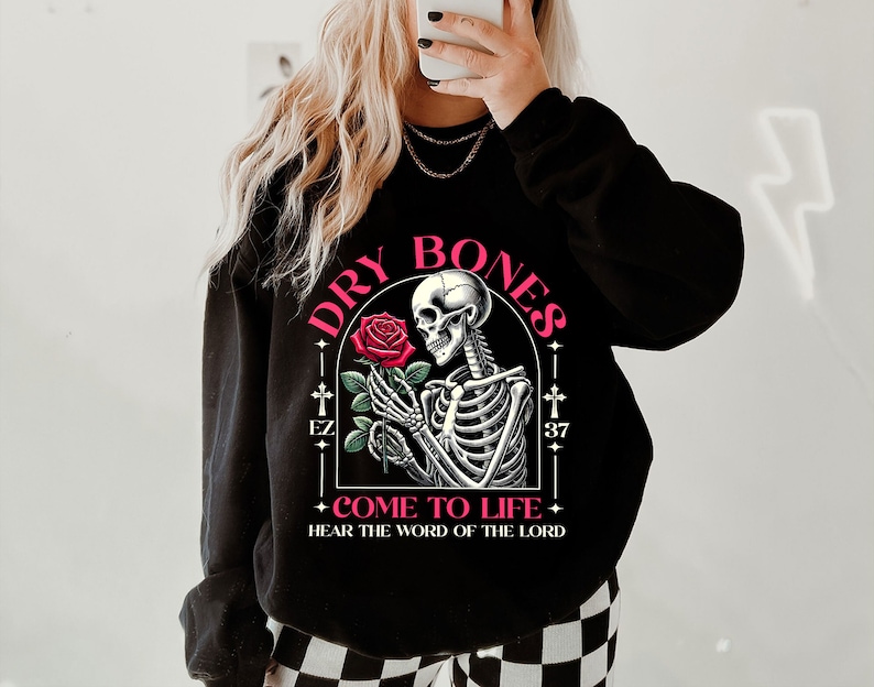 May include: Black sweatshirt with a graphic of a skeleton holding a red rose. The graphic is surrounded by a black frame with the text "DRY BONES" in pink letters and "COME TO LIFE - HEAR THE WORD OF THE LORD" in smaller pink letters.