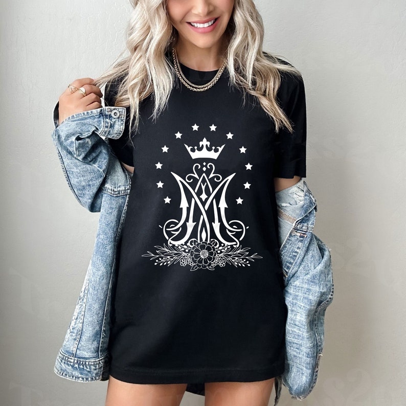 May include: Black t-shirt featuring a white graphic of a crown above the letters "M" and "A" surrounded by stars and floral accents.