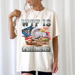 May include: Cream-colored t-shirt featuring a graphic design with a bald eagle, American flag, and a tank. The text "WTF IS A KILOMETER" is displayed in a bold, metallic font.
