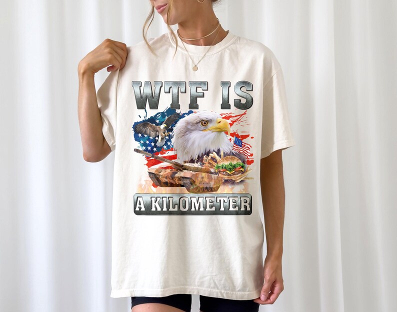 May include: Cream-colored t-shirt featuring a graphic design with a bald eagle, American flag, and a tank. The text "WTF IS A KILOMETER" is displayed in a bold, metallic font.