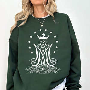 May include: A dark green sweatshirt featuring a white graphic design of a crown, stars, and a stylised letter "M" with floral accents.