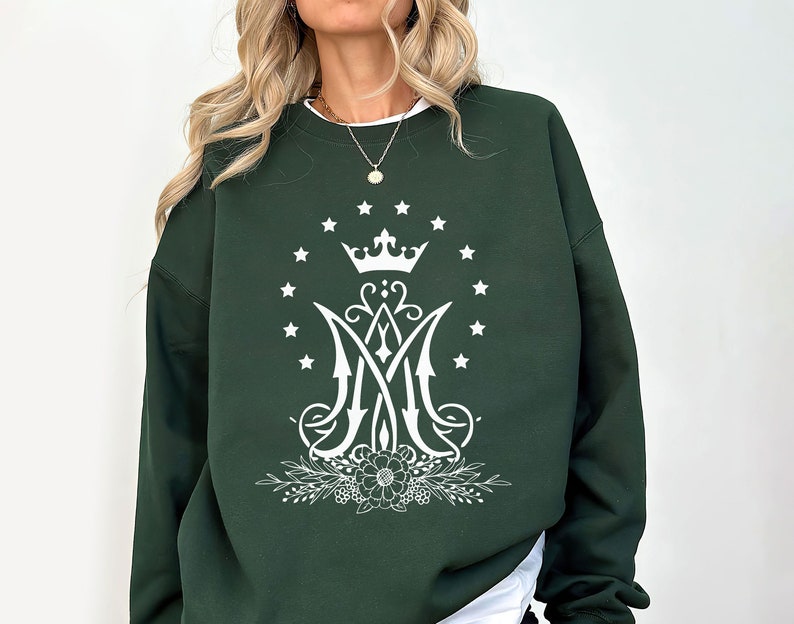 May include: A dark green sweatshirt featuring a white graphic design of a crown, stars, and a stylised letter "M" with floral accents.
