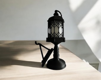 May include: Black metal candle lantern with a decorative design and a white candle inside. The lantern is on a black metal stand with a base.