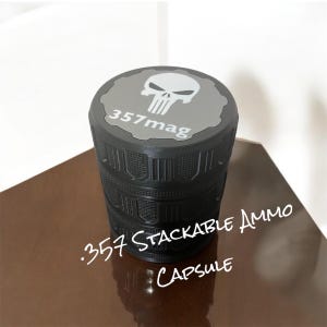 May include: Black stackable ammo capsule with a grey top featuring a white skull logo and the text "357mag". The capsule has a textured design and the words ".357 Stackable Ammo Capsule" are written on the side.