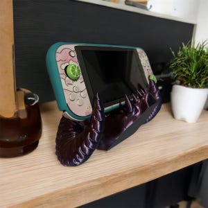 May include: A purple and black 3D printed stand shaped like a pair of demon horns holds a teal and white Nintendo Switch console.