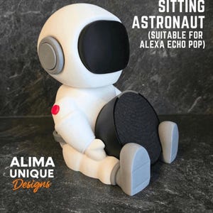Puede incluir: Una figura de astronauta blanco sentado, con visera negra y detalles grises. La figura está diseñada para sostener un altavoz Alexa Echo Pop. Los textos "SITTING ASTRONAUT" y "ALIMA UNIQUE Designs" son visibles.