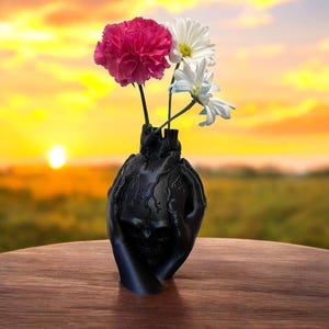 Puede incluir: Un jarrón decorativo negro con forma de corazón y manos, que sostiene flores rosas y blancas. El jarrón tiene un diseño de calavera y está sobre una superficie de madera. El fondo es una puesta de sol.