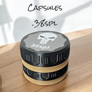 Może przedstawiać: Stosowane kapsułki na amunicję w kolorze czarnym i beżowym z tekstem ".38SPL" i "CAPSULES". Wierzch kapsułki ma szary wzór z grafiką czaszki i napisem "38spl". Kapsułki są ułożone na drewnianej powierzchni.