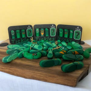 May include: A set of green pickle finger puppets with different facial expressions. The puppets are packaged in three black boxes with the words "Tickles Tickles" and "4 Pack" printed on them.