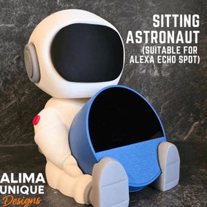 Puede incluir: Una figura de astronauta blanca y gris que sostiene un dispositivo azul y negro, diseñado para el Alexa Echo Spot. La figura tiene una visera negra y detalles grises. El texto en la imagen dice "SITTING ASTRONAUT" y "ALIMA UNIQUE Designs".