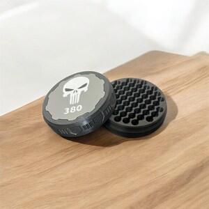 May include: A gray and black round grinder with a skull logo and the number 380 printed on the lid. The grinder is open, revealing the grinding teeth. The grinder is on a wooden surface.