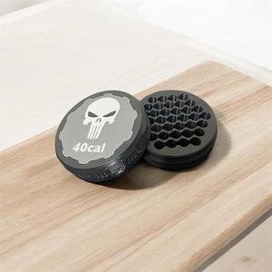 May include: Two-piece, dark gray ashtray set with a skull graphic and the text "40cal" on the lid. The ashtray has a honeycomb-patterned interior. The set is displayed on a light wood surface.