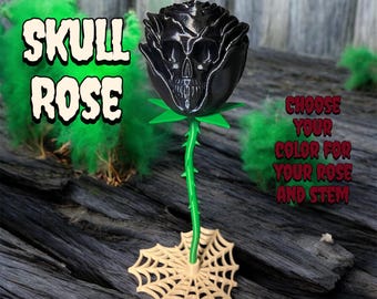 May include: A black skull rose with a green stem and leaves, and a beige spiderweb base. The text "SKULL ROSE" is in a green and white font. The text "CHOOSE YOUR COLOR FOR YOUR ROSE AND STEM" is in a red and white font.