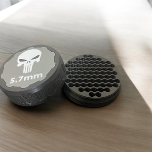 May include: Two-piece, gray metal grinder with a skull logo and the text "5.7mm" on the lid. The other piece has a honeycomb-like pattern. The grinder is designed for a specific purpose, likely related to herbs or spices.