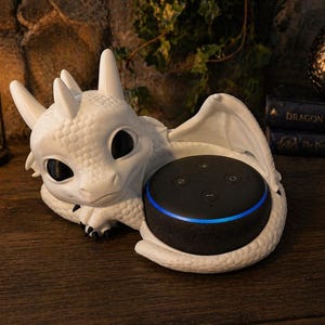 May include: A white dragon-shaped speaker holder containing a black smart speaker with a blue light. The dragon has large black eyes and detailed scales. The speaker is on a wooden surface.