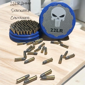 May include: A blue stackable container filled with .22LR ammunition, with additional rounds scattered on a wooden surface. The container lid features a skull graphic and the text "22LR". The text "22LR Ammo Stackable Container" is also visible.