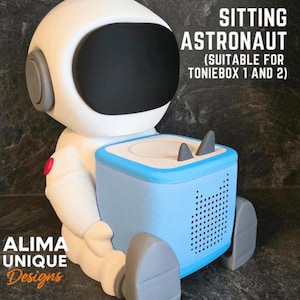 May include: A white astronaut figure with a black visor and gray accents, holding a light blue and white rectangular device. The text "SITTING ASTRONAUT (SUITABLE FOR TONIEBOX 1 AND 2)" is visible. The figure is labeled "ALIMA UNIQUE Designs".