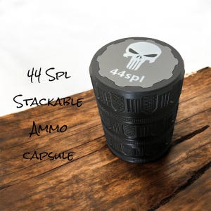 May include: A black stackable ammo capsule with a gray top featuring a white skull design and the text "44spl". The text "44 Spl Stackable Ammo Capsule" is written on the left side of the image. The capsule is on a wooden surface.