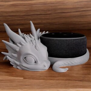 May include: A gray 3D printed dragon head with a curled tail wraps around a black cylindrical smart speaker.