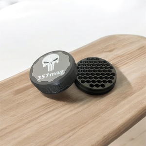 May include: A gray and black grinder with a skull logo and the text "357mag". The grinder is round with a textured grip and a separate piece with a honeycomb pattern. The items are on a wooden surface.