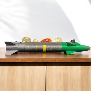 May include: A green and gray missile-shaped desk organizer with the text "U.S.M.C." on the side. The organizer has a slot for holding small items, such as coins or pins. There are several coins and pins in the slot.