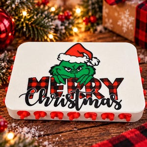 Könnte beinhalten: Ein rechteckiges weißes Kunststoffgerät mit einer Reihe kleiner roter Vorsprünge an der Unterkante. Die Oberseite zeigt eine Grafik einer grünen Figur mit roter Weihnachtsmütze über dem rot-schwarz karierten Schriftzug MERRY CHRISTMAS.