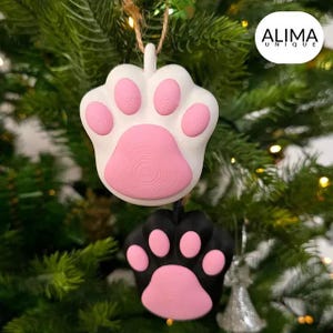 以下が含まれることがあります： ピンクと黒の肉球の形をしたクリスマスオーナメント2つ。オーナメントはクリスマスツリーに飾られています。