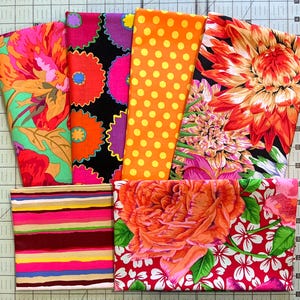 May include: A collection of colorful fabric swatches with floral, polka dot, and striped patterns. The fabrics feature vibrant hues of orange, pink, green, and purple. The fabrics are arranged on a cutting mat.