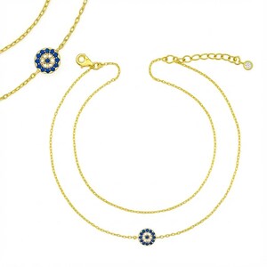Może przedstawiać: Złoty, warstwowy naszyjnik z dwoma łańcuszkami. Każdy łańcuszek posiada małą, okrągłą zawieszkę z niebiesko-białym wzorem. Naszyjnik posiada zapięcie na karabińczyk i regulowany łańcuszek.