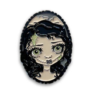 May include: An oval-shaped enamel pin featuring a stylized zombie girl with black hair, green eyes, and stitched facial features. The pin has a black border and a light beige background.
