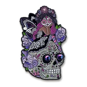Puede incluir: Pin decorativo con un diseño de calavera de azúcar con un hada. La calavera es plateada con detalles morados y purpurina brillante. El hada tiene pelo morado, una corona floral y alas de mariposa. El pin está rodeado de flores moradas.