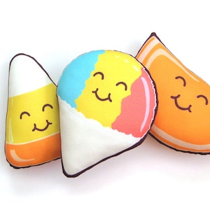 Puede incluir: Tres peluches de comida: un caramelo de ma&iacute;z, un cono de nieve y una rodaja de naranja. Cada uno tiene una cara sonriente. El caramelo de ma&iacute;z es amarillo, naranja y blanco. El cono de nieve es azul, amarillo y rosa. La rodaja de naranja es naranja.