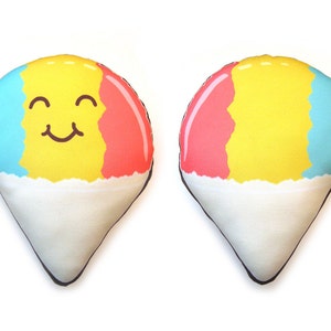 Puede incluir: Dos cojines de felpa con forma de cono de nieve. Uno tiene una cara sonriente, con secciones azules, amarillas y rojas sobre una base blanca. El otro coj&iacute;n tiene el mismo patr&oacute;n de color sin cara. Ambos tienen un contorno negro.